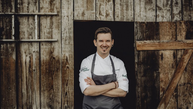 A smiling man in chef's clothes stands in front of a wooden wall.