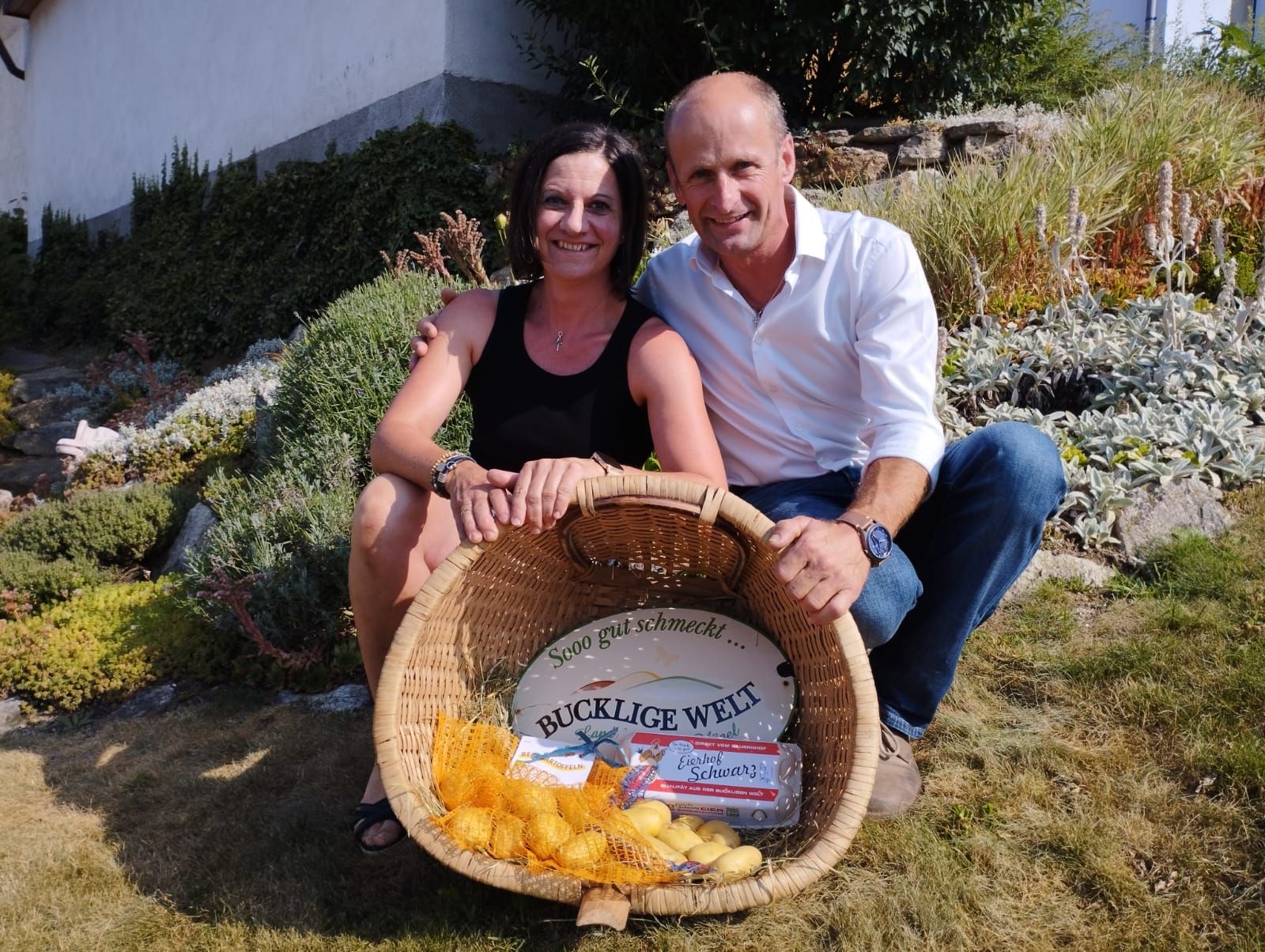 A woman and a man sit smiling in a garden, holding a basket of potatoes and eggs.