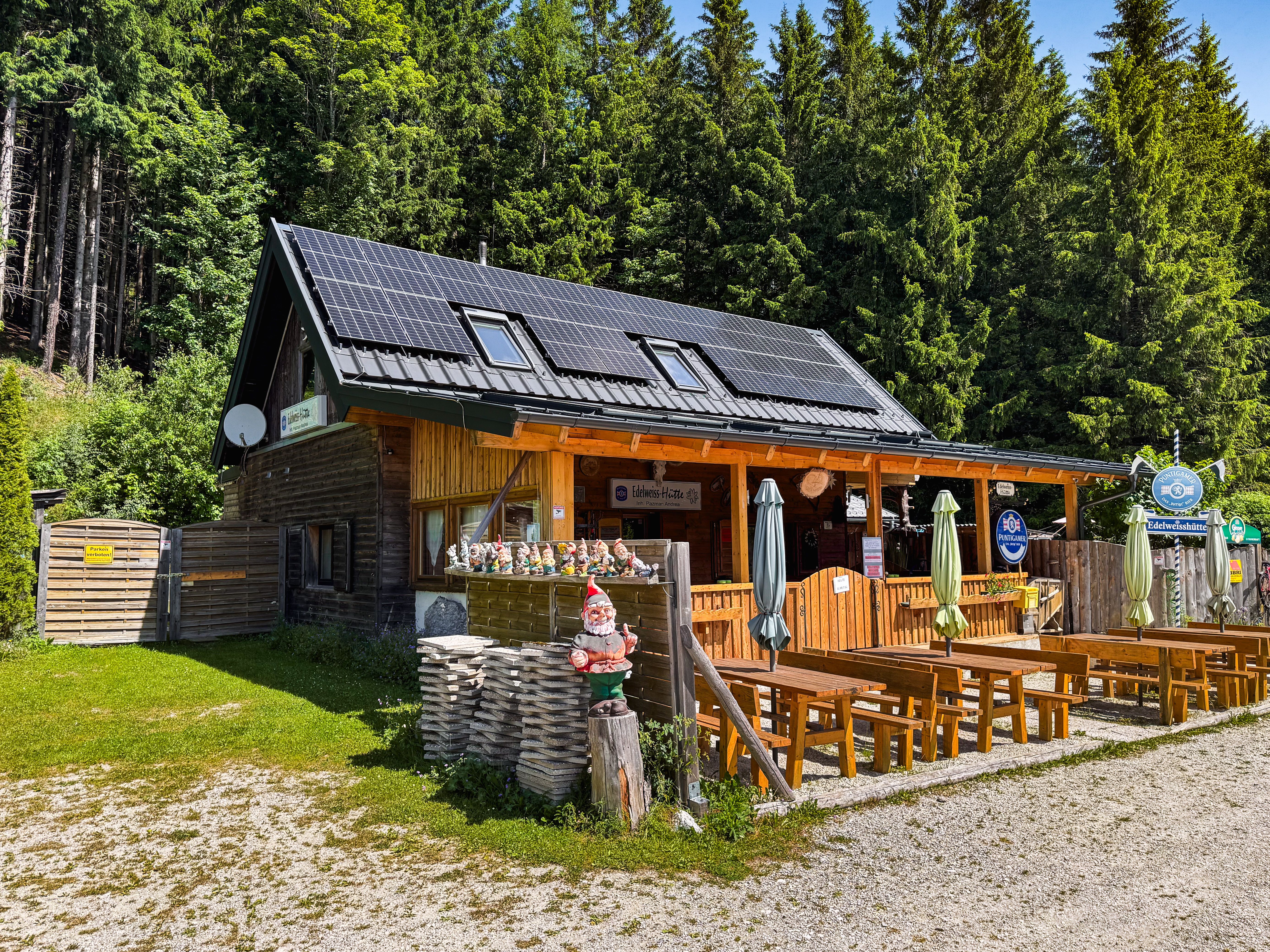 A mountain hut with solar panels on the roof, surrounded by trees. There are wooden benches and a garden gnome in front of the hut.