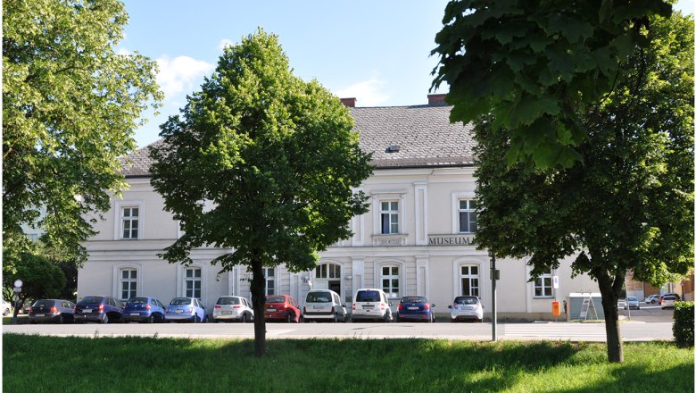 A white building with the inscription 'Museum', surrounded by trees and parked cars.