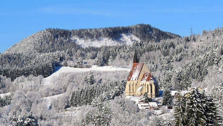 Winter landscape with a church in front of a wooded hill.