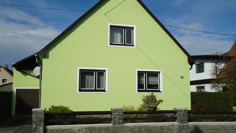A light green house with a pitched roof and two windows, surrounded by a low stone fence.