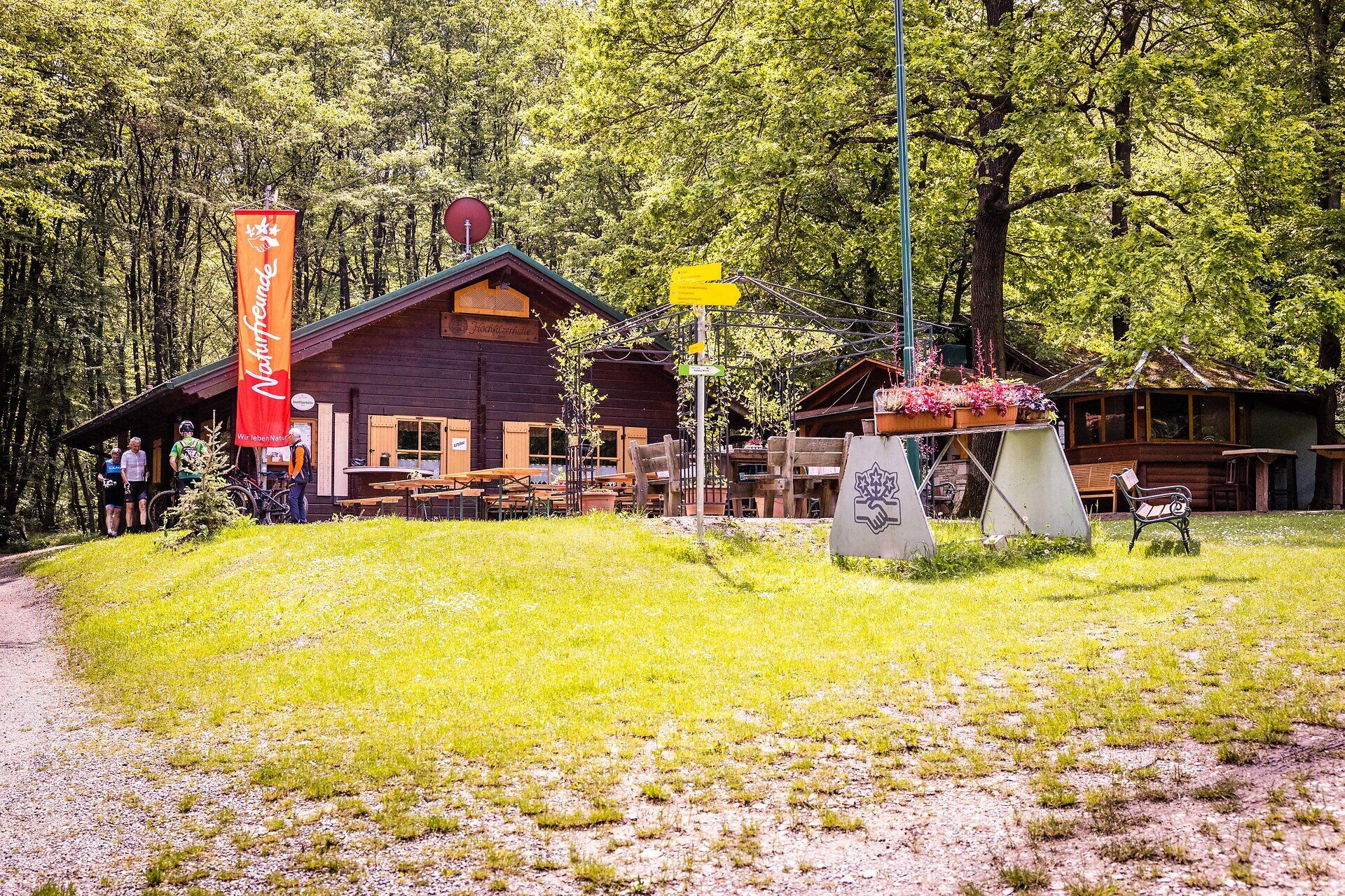 Nature lovers' wooden hut with tables, benches and people in front of it. 