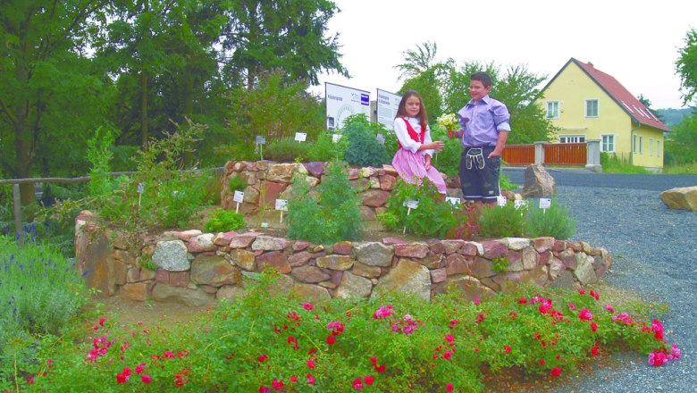 Two children stand in a herb spiral made of stones, surrounded by flowering plants and a yellow house in the background.
