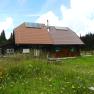 A mountain hut with solar panels on the roof, surrounded by a flowering meadow and forest in the background.