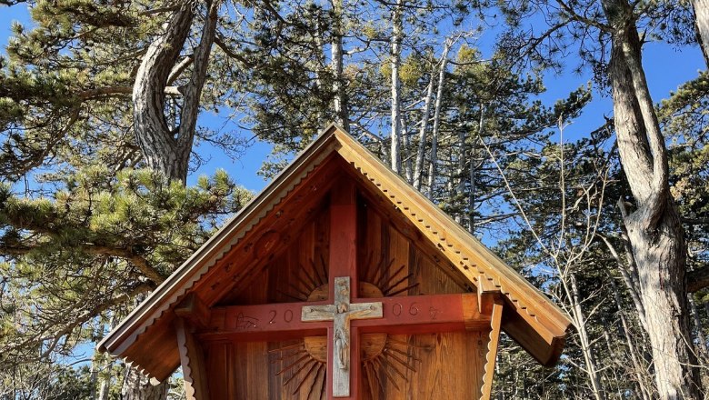 Wooden cross with roof in a forest, surrounded by trees and blue sky.