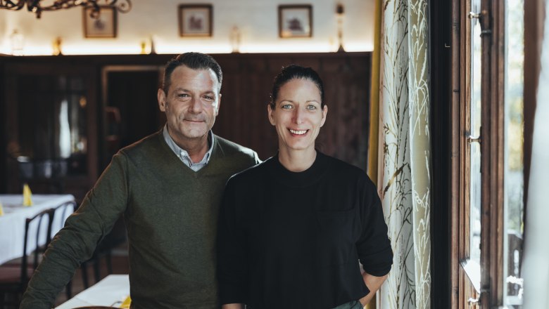 A man and a woman stand smiling in a cozy restaurant.