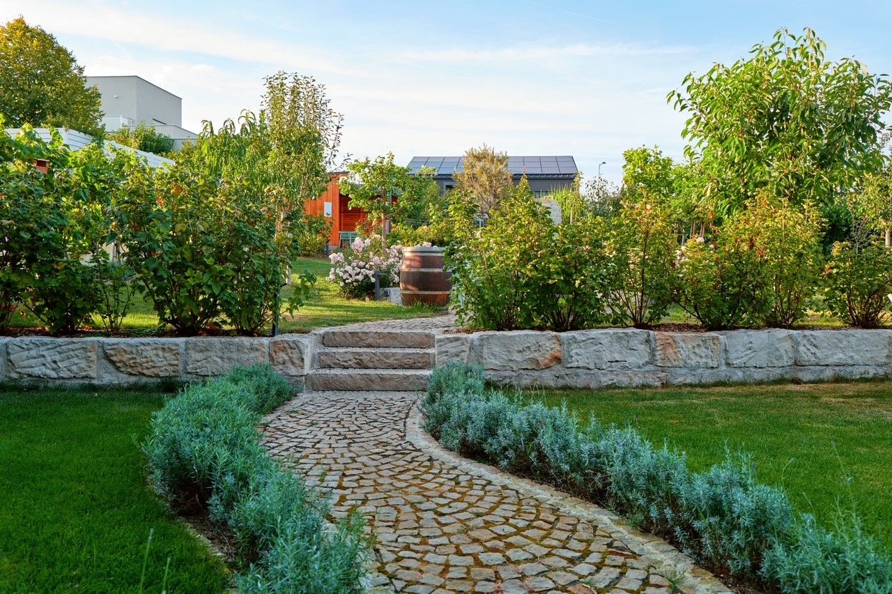 Well-kept garden with stone path, bushes and trees, in the background a building with solar panels.