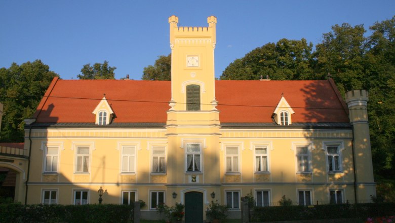 Yellow castle with red roof and tower, surrounded by trees.