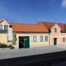 A long, single-storey house with a yellow and pink façade, green shutters and a red tiled roof under a blue sky.