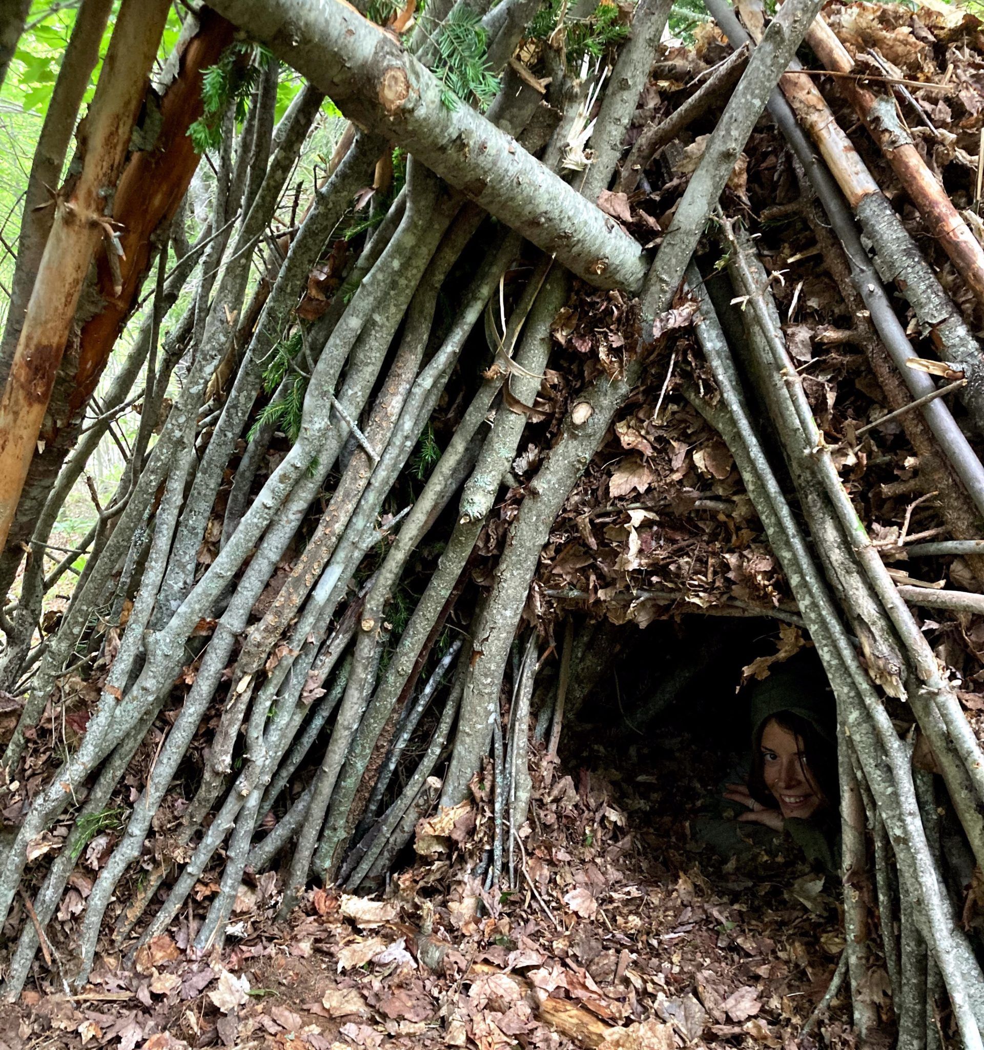 A person peers out of a self-made shelter made of branches and leaves in the forest.