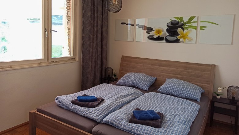 Bedroom with double bed, blue bed linen, picture with stones and flowers on the wall, window and bedside lamp.