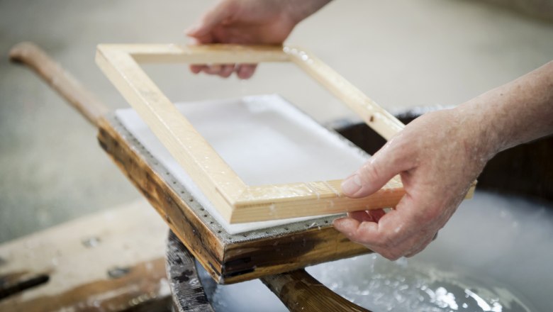Close-up of hands holding a wooden frame over a paper pulp in a traditional paper mill.