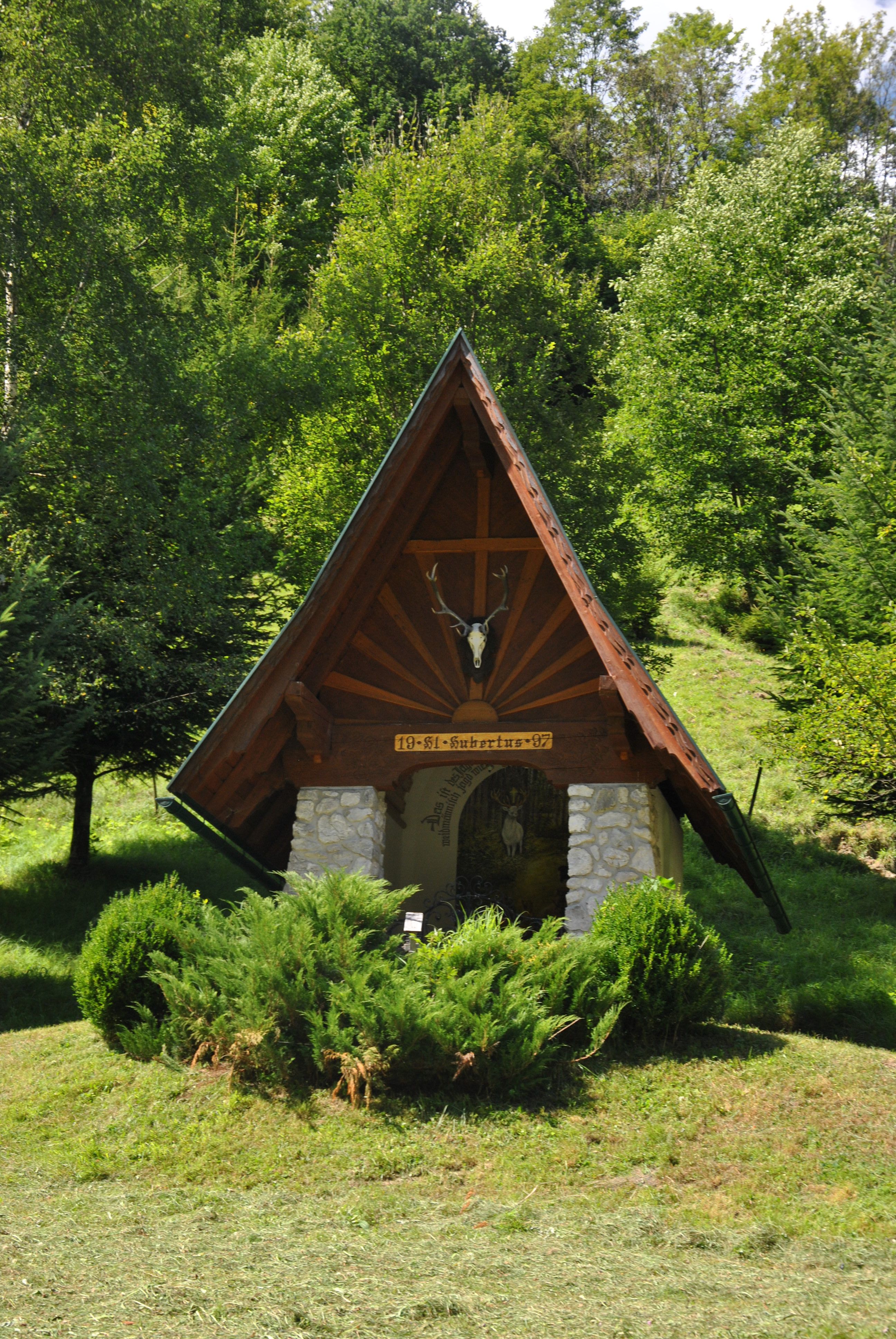 A small chapel with a pointed roof and deer antlers, surrounded by trees and bushes.