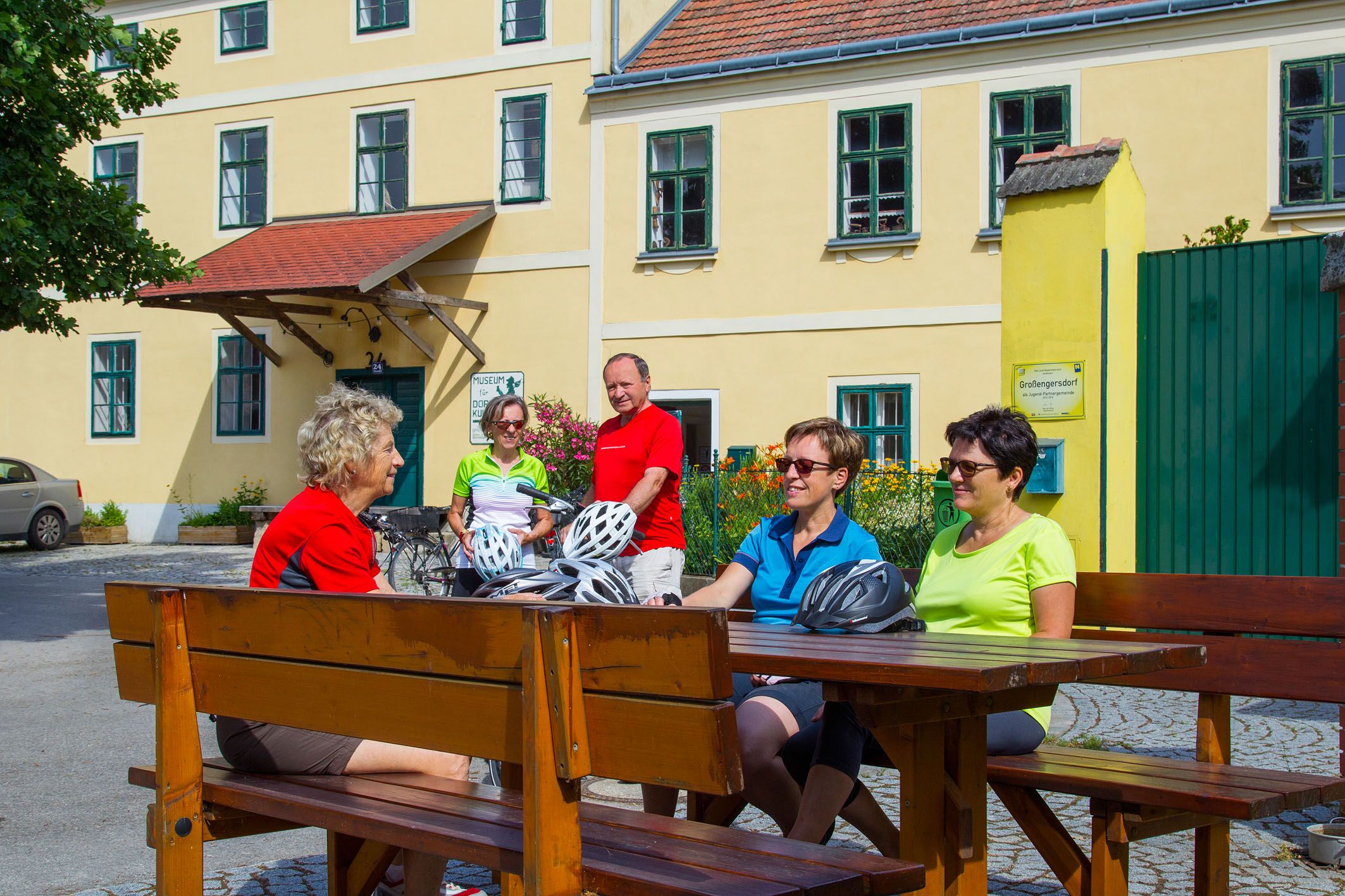 Group of cyclists sitting at a wooden table in front of a yellow building.