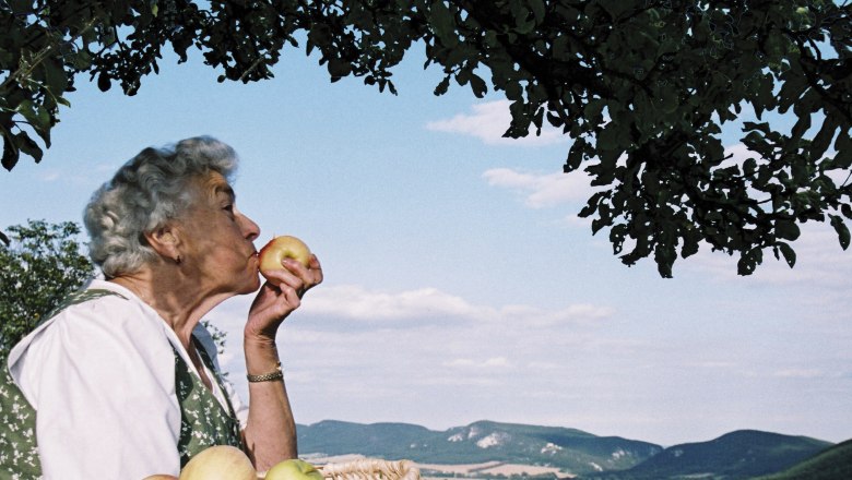 Elderly woman with a basket full of apples kissing an apple in front of a landscape with hills and blue sky.