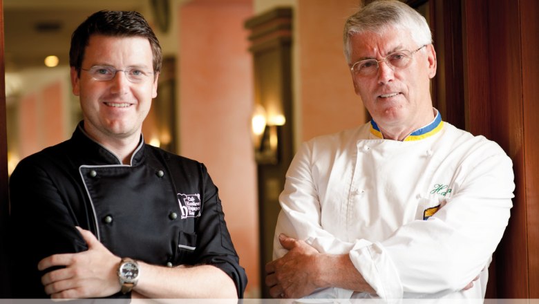 Two men in pastry chef uniforms stand next to each other and smile at the camera.