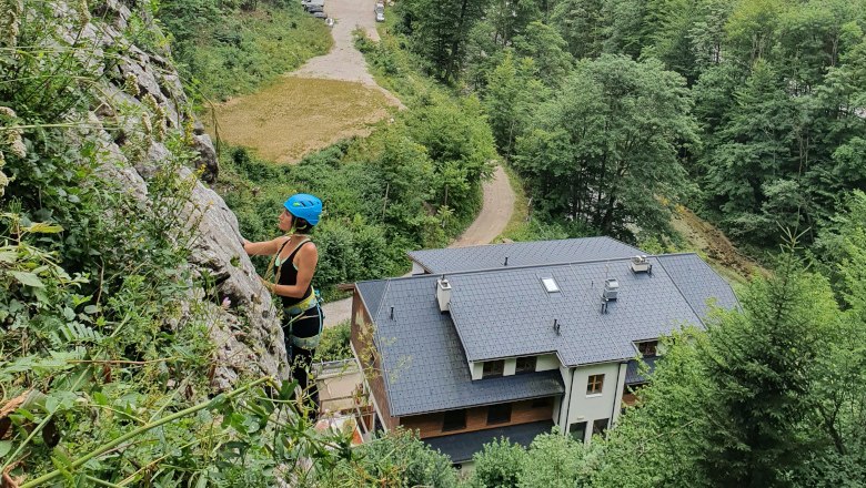 Person climbing on a rock face with helmet and climbing equipment, buildings and forest in the background.