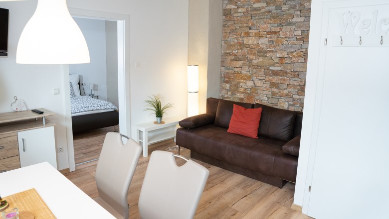 Modern living room with brown sofa, stone wall and view into the bedroom.