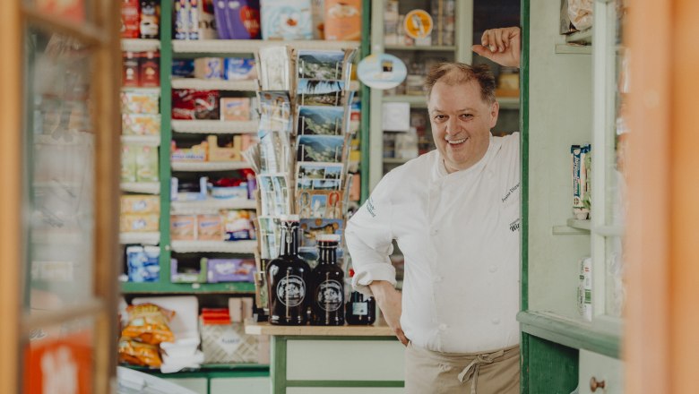 A man in chef's clothes stands smiling in an old store with shelves full of products.