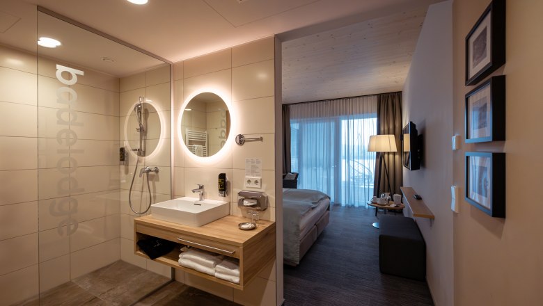 Modern hotel room with open bathroom, washbasin, mirror and shower. In the background a bed and a window with curtains.