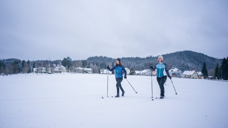 Two people cross-country skiing on a snow-covered surface in front of a village and wooded hills.