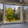 View from an open window of a green garden and a historic building with a tower.