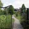 An open wrought-iron gate leads to a paved path that leads through a green garden to a house in the background.