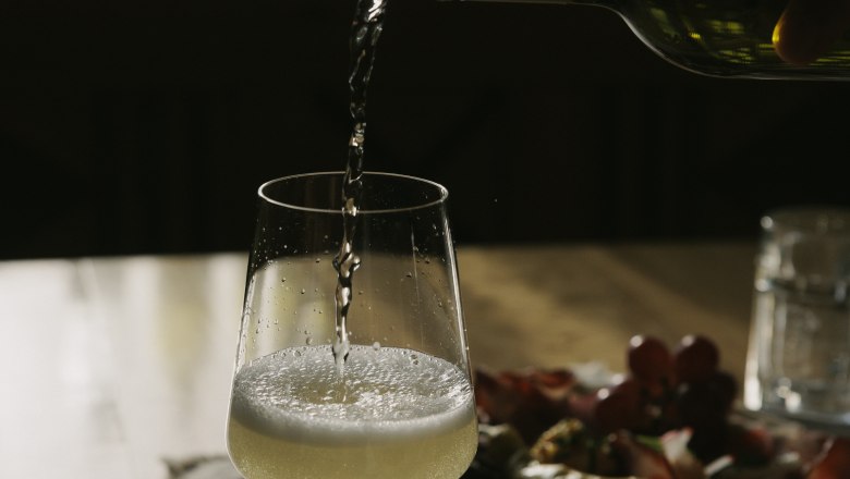 A glass of white wine is poured on a table, with a plate of snacks in the background.