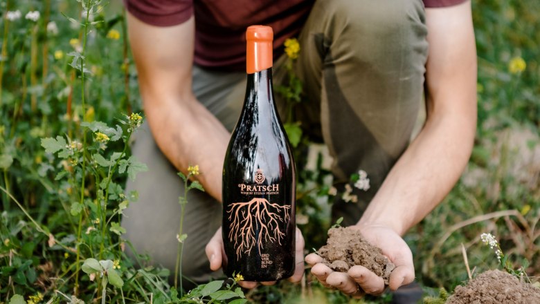 A person holds a bottle of Pratsch wine and a handful of soil in a green field.