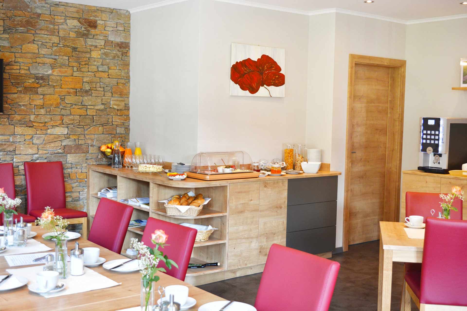 A breakfast buffet in a cozy room with red chairs, wooden furniture and a stone wall. The buffet includes croissants, muesli and orange juice.