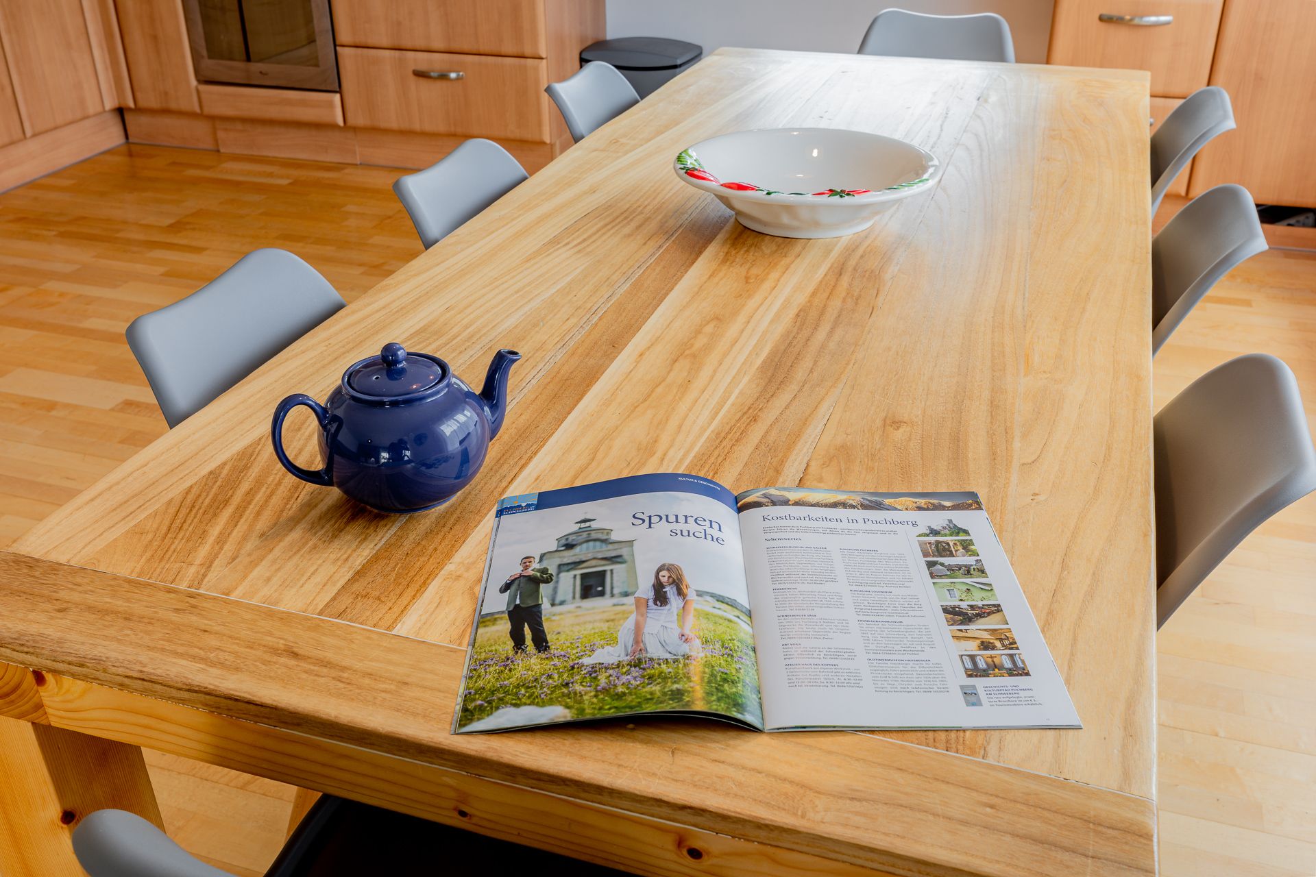 A wooden table with a blue teapot, a bowl and a magazine in a kitchen.