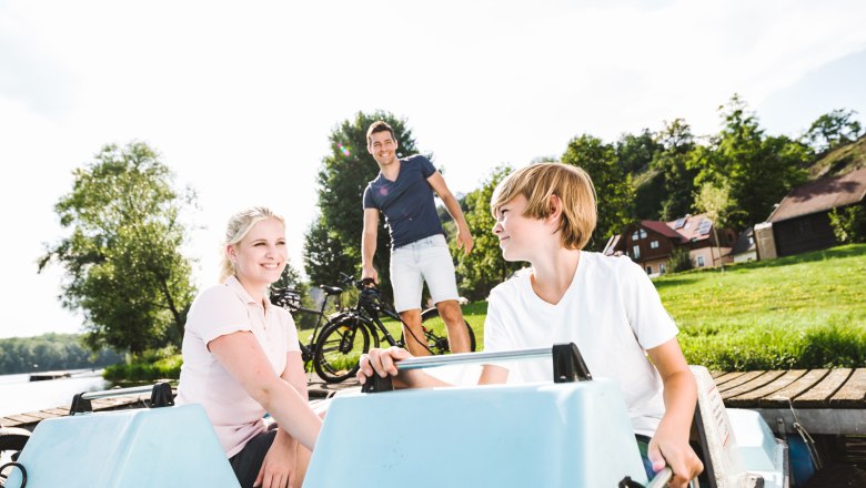 Two children in a pedal boat, a man with a bicycle in the background, green meadow and trees.