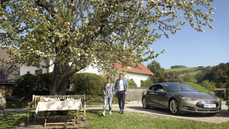 A couple walks next to a parked electric car under a blossoming tree in a rural garden.