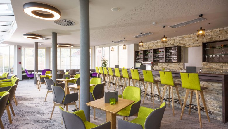 Modern café with green and gray chairs, wooden tables and a long bar with a stone wall.