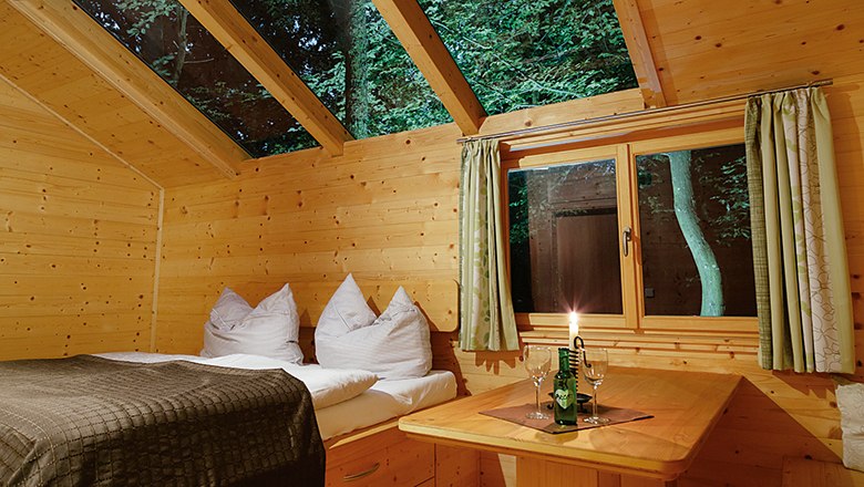 Interior view of a wooden hut with bed, table and candle, trees are visible through the window and skylight.
