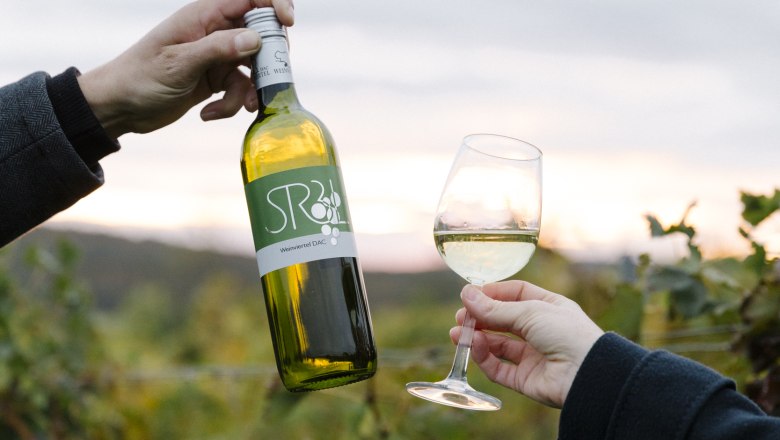Two hands hold a bottle of wine and a glass of white wine in front of a vineyard.