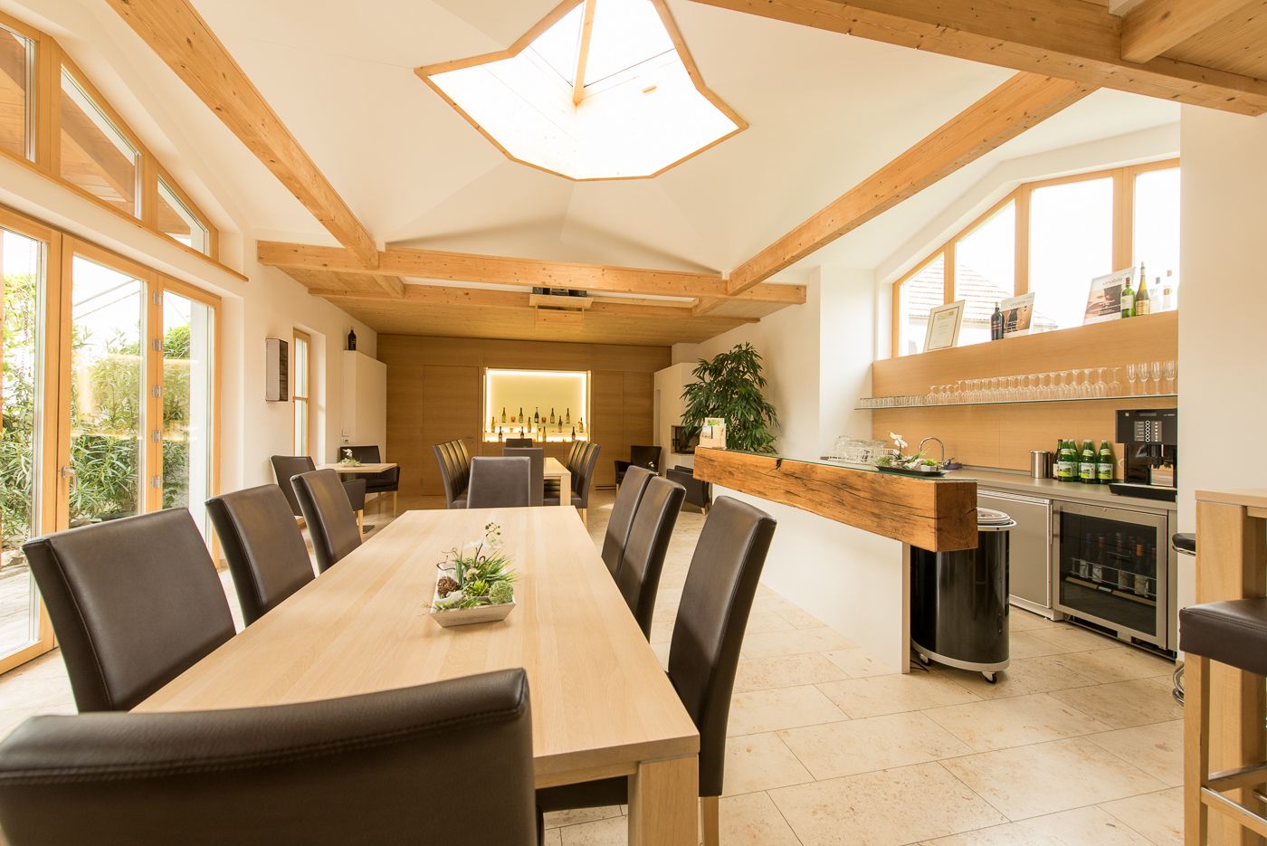 Interior view of a modern tasting room with wooden tables and chairs, large windows and a bar with bottles of wine.