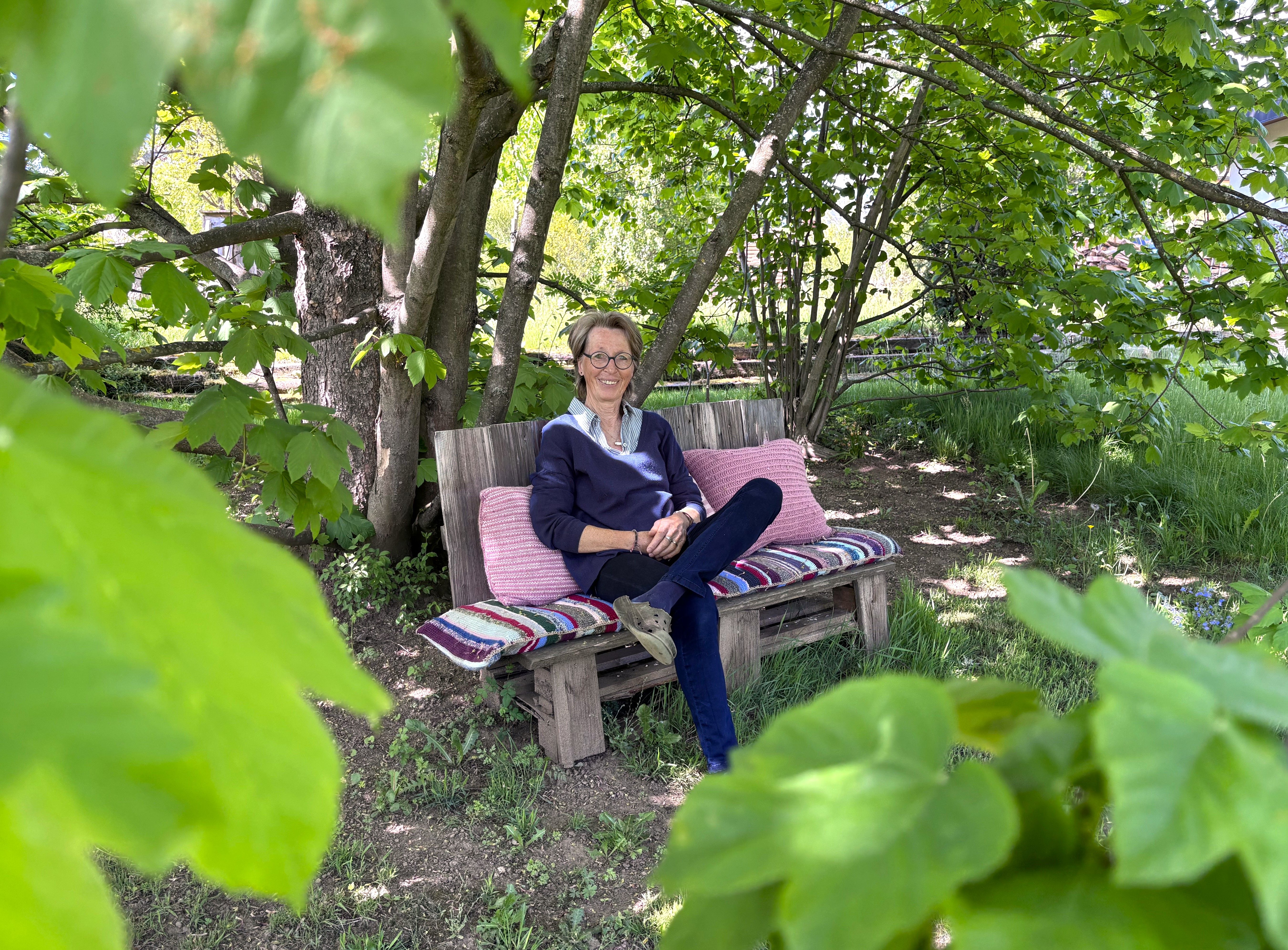 A person is sitting on a bench in the garden, surrounded by green leaves and trees.