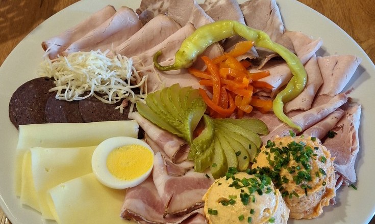 A snack plate with various types of sausage and cheese, egg, peppers, gherkins and spread on a wooden table.