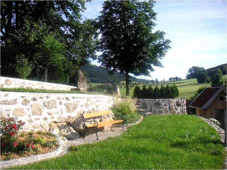 A garden with a stone wall, bench and trees in the background.