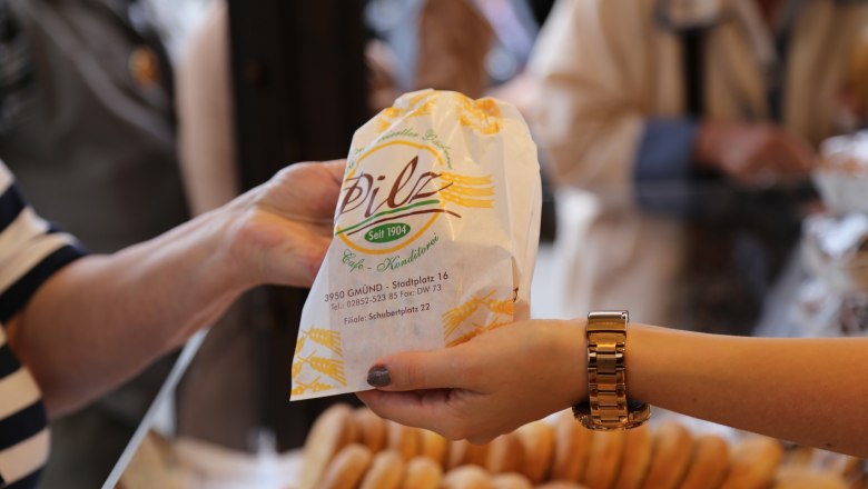 Two hands hand over a paper bag from the Pilz bakery-pastry shop with pastries in the background.