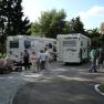 People stand and sit next to two mobile homes on an outdoor pitch.