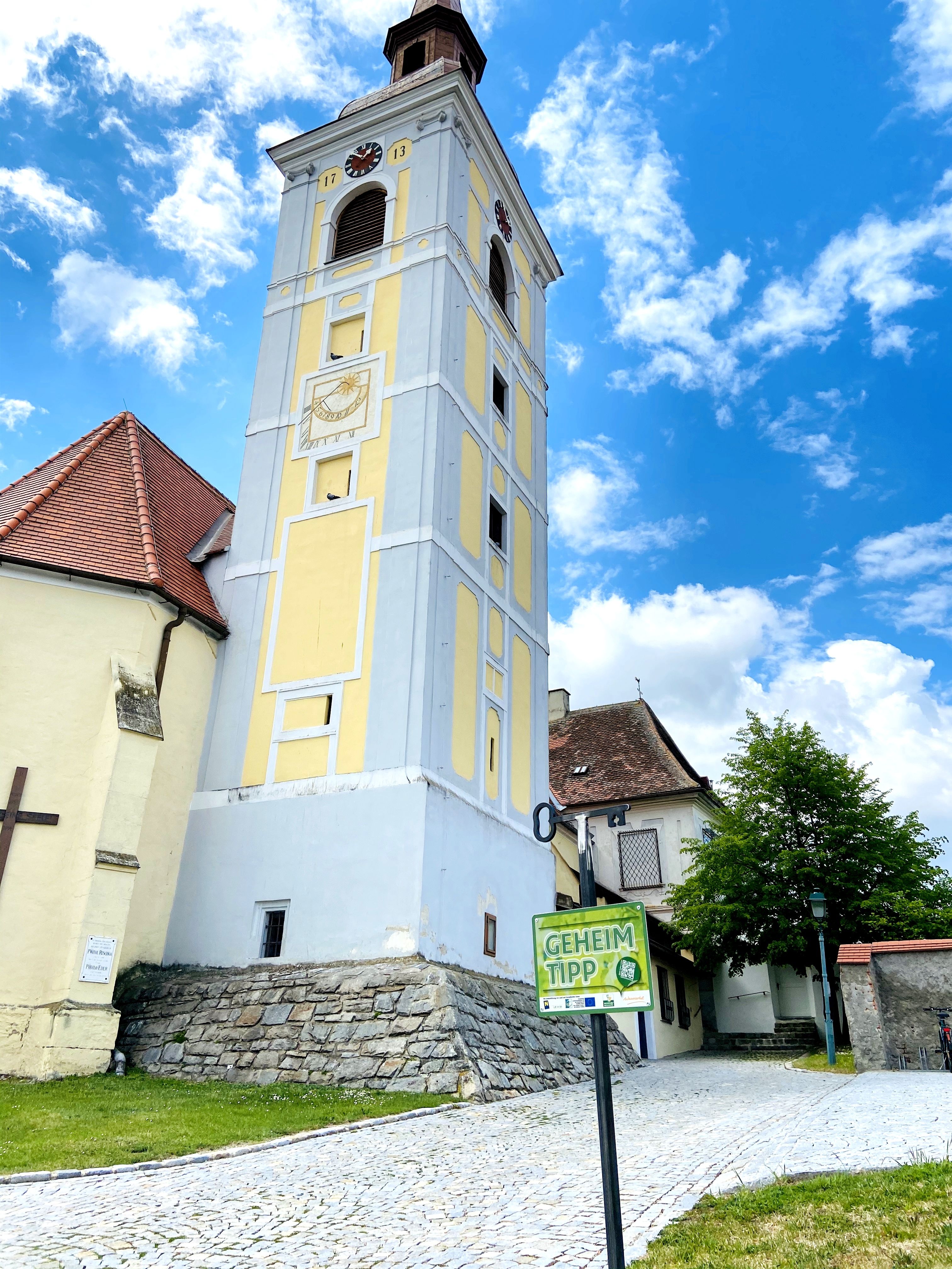 Leaning tower in Waitzendorf with yellow facade and clock, surrounded by blue sky and clouds.