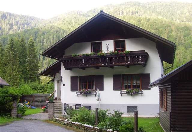 A traditional vacation home with a wooden balcony and flower boxes, surrounded by green trees and mountains.