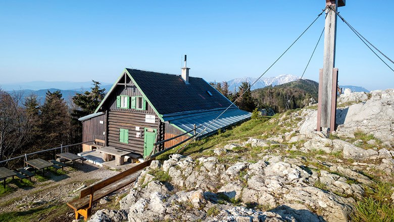 A mountain hut with green shutters and a summit cross in the foreground, surrounded by rocks and trees.