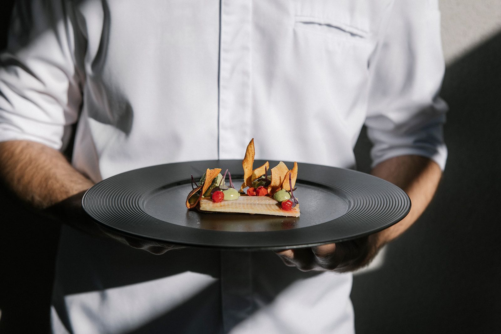 A person in a white shirt holds a plate with an elaborately prepared gourmet dish.
