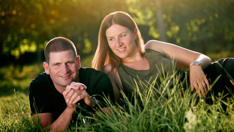 A man and a woman lie relaxed on the grass, surrounded by green nature and sunlight.