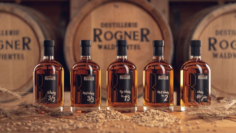 Five bottles of whisky from the Rogner distillery in front of wooden barrels.