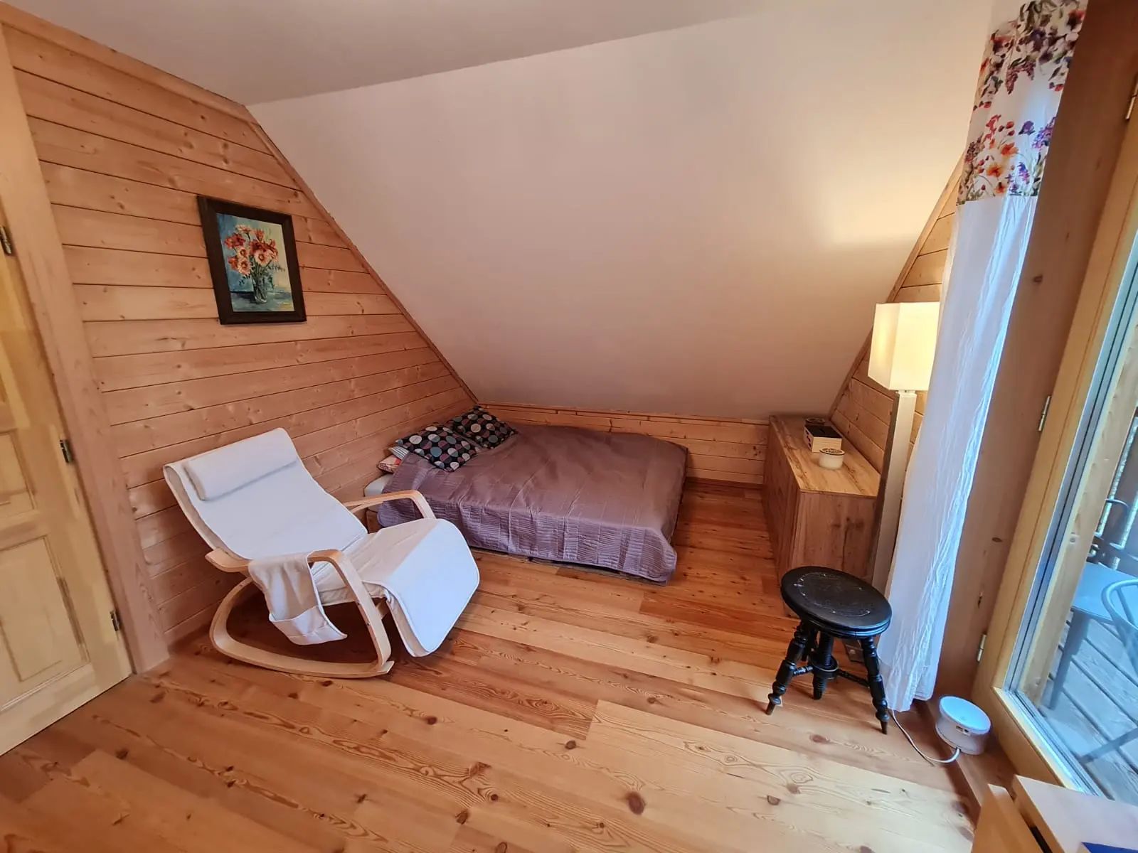 Cozy room with wooden walls, a bed, a white rocking chair and a small table.